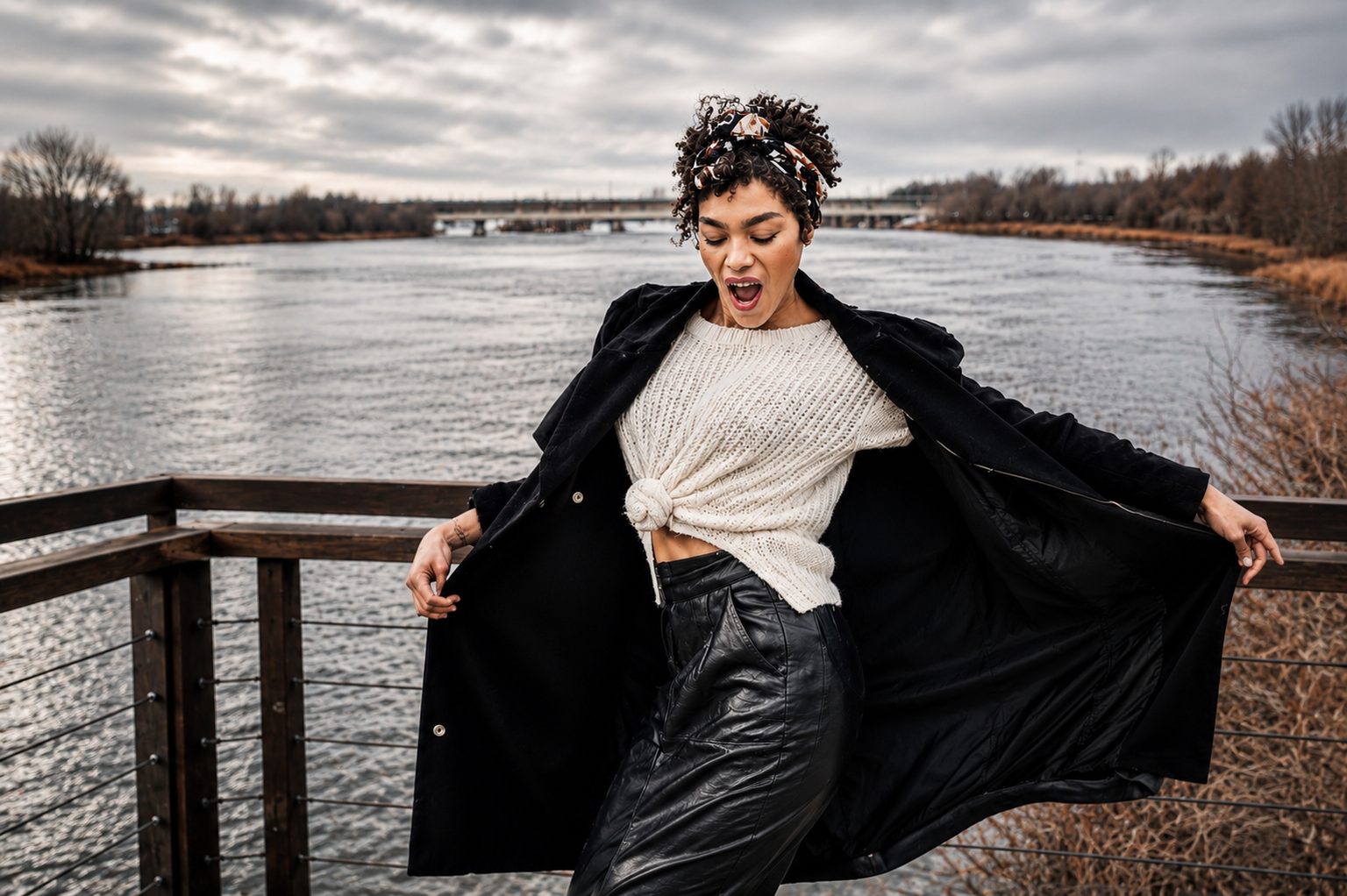 Dramatic editorial portrait on a lakeside boardwalk, coat caught mid-movement